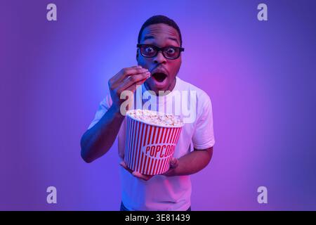 Homme noir choqué portant une chemise blanche et des lunettes de soleil mangeant du pop-corn tout en se tenant debout sur fond violet néon en studio Banque D'Images