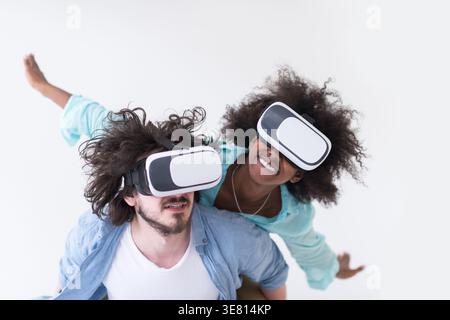 Couple noir et caucasien portant des casques VR ayant un plaisir piggyback équitation et expérimentant la réalité virtuelle sur fond blanc en studio Banque D'Images