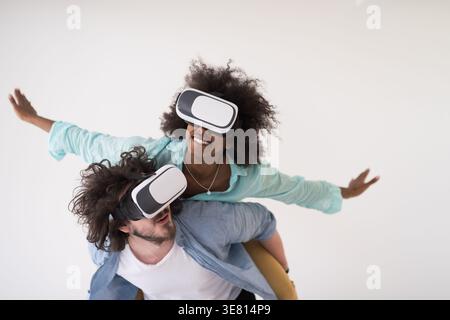Couple noir et caucasien portant des casques VR ayant un plaisir piggyback équitation et expérimentant la réalité virtuelle sur fond blanc en studio Banque D'Images