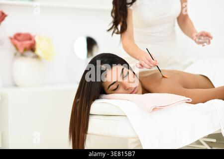 Gardienne du spa féminine du moyen-Orient appliquant un traitement de massage du dos à l'aide d'une brosse à huile à femme de race mixte avec les yeux fermés couchés sur le lit dans le puits... Banque D'Images
