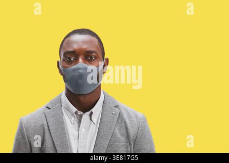 Homme d'affaires afro-américain en costume gris portant un masque facial debout devant un fond jaune dans le studio Banque D'Images