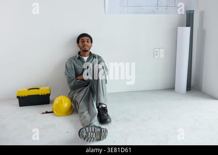 Constructeur masculin noir fatigué assis avec les yeux fermés et penché en arrière sur le mur prenant une sieste pendant la pause déjeuner Banque D'Images