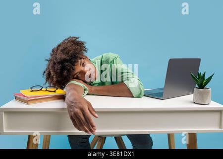 Étudiant noir épuisé avec les cheveux bouclés penchant le visage sur son bras dormant sur la table avec livre et ordinateur portable devant le fond bleu dans le studio Banque D'Images