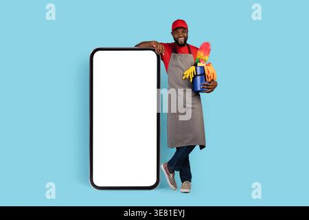 Noir nettoyant masculin chapeau de port et tablier tenant panier d'outils de nettoyage debout et penché avec les jambes croisées sur grand smartphone contre bac bleu... Banque D'Images