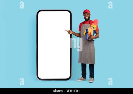 Chapeau de port de nettoyant masculin noir et tablier pointant vers le grand smartphone tenant le panier d'outils de nettoyage debout sur fond bleu dans le studio Banque D'Images