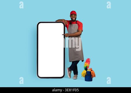 Casquette et tablier noir nettoyant masculin pointant sur le grand smartphone debout avec les jambes croisées sur fond bleu en studio Banque D'Images