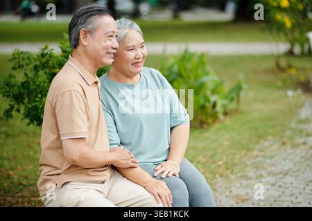 Couple aîné asiatique regardant loin assis ensemble dans le parc de la ville pendant la journée Banque D'Images