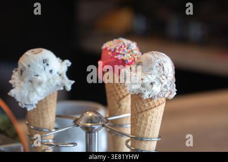 Trois cônes de crème glacée avec différentes saveurs et garnitures sur support en métal contre fond de café, lumière douce et faible profondeur de champ. Nourriture Banque D'Images