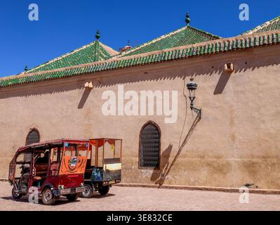 Façade latérale du bâtiment historique de la Madrasa Ben Youssef avec deux tuk-tuks garés à l'extérieur par une journée d'été ensoleillée à Marrakech, Maroc Banque D'Images
