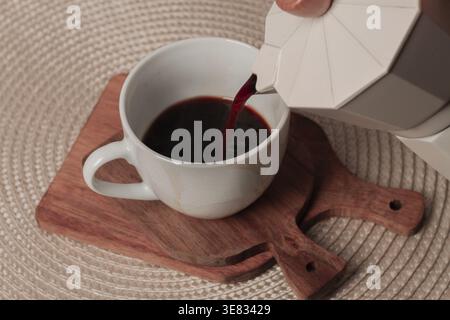 Le café fraîchement infusé est soigneusement versé d'une cafetière de cuisinière dans une petite tasse blanche, reposant sur une planche à découper en bois, créant un coz Banque D'Images