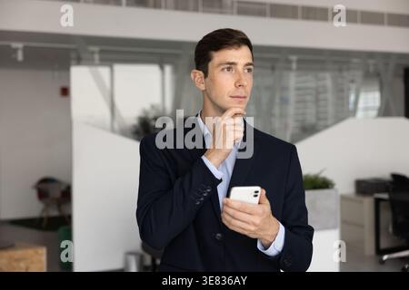 Homme d'affaires pensif habillé en costume se tient dans le bureau avec smartphone Banque D'Images