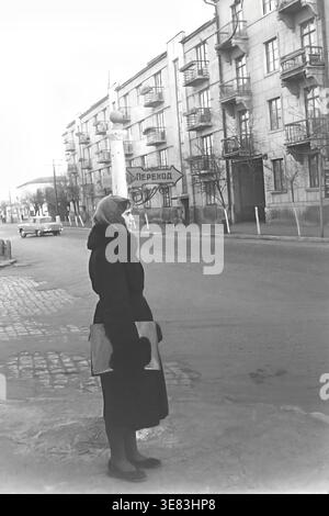 Slaviansk, années 1950 Une vue urbaine historique captivante d'Ulitsa Karla Marksa (aujourd'hui Tsentralna) avec une femme vêtue d'un manteau d'hiver sombre et d'un foulard debout près d'une route pavée. Elle tient un grand dossier, regardant soigneusement devant. En arrière-plan, un bâtiment résidentiel classique de plusieurs étages avec balcons, un panneau de passage pour piétons et une voiture soviétique d'époque créent une scène de rue vivante. Cette authentique photographie en noir et blanc préserve le patrimoine architectural unique et la vie tranquille dans la rue du Donbass paisible. Banque D'Images