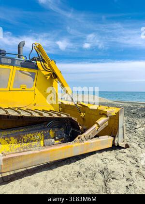Un bulldozer jaune travaille sur la plage, déplaçant le sable près de l'eau. Le ciel clair et l'océan sont visibles en arrière-plan, montrant l'activité le long des s. Banque D'Images