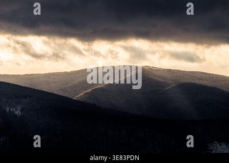 Pentes montagneuses hivernales couvertes de neige et de gel au coucher du soleil, rayons de lumière dorée illuminant les vallées forestières brumeuses, paysages montagneux de mauvaise humeur Banque D'Images
