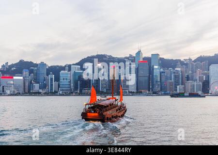 Jonque traditionnelle en bois avec des voiles rouges contre Hong Kong skyline pendant le coucher du soleil, scène urbaine emblématique Banque D'Images