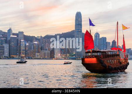 Jonque traditionnelle en bois avec des voiles rouges contre Hong Kong skyline pendant le coucher du soleil, scène urbaine emblématique Banque D'Images