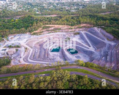 Vue aérienne d'une carrière à ciel ouvert active montrant des rochers en terrasses, des étangs turquoises, une autoroute, une forêt luxuriante et des logements lointains de banlieue à Brisbane. Banque D'Images