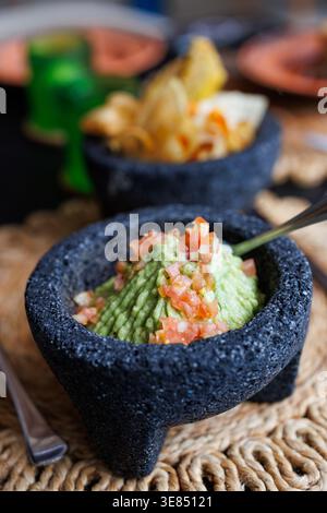 Gros plan de guacamole frais recouvert de pico de gallo dans un molcajete en pierre volcanique noire, avec des copeaux de tortilla en arrière-plan, cadre de table rustique Banque D'Images