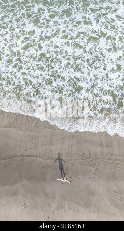 Vue aérienne des vagues de l'océan qui se jettent sur la plage de sable avec une personne seule projetant une longue ombre, surf texturé et rivage, voyage d'été minimal Banque D'Images