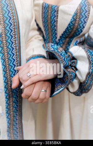 Un plan vertical d'un couple habillé en tenue traditionnelle slave ukrainienne avec broderie bleue, leurs mains ornées d'anneaux de mariage Banque D'Images