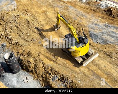 Vue aérienne d'une pelle à chenilles jaune creusant et nivelant du sol pour une tranchée de fondation sur un chantier de construction actif, montrant des chenilles de terrassement Banque D'Images