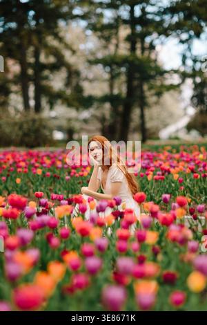 Jeune femme sereine aux cheveux rouges dans une robe blanche reposant parmi les tulipes vibrantes dans un parc de printemps, les arbres doux derrière, l'humeur paisible et la beauté naturelle Banque D'Images