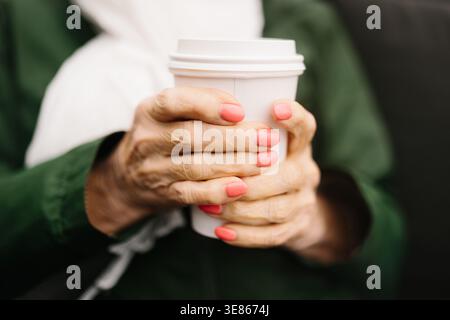 Gros plan des mains d’une femme avec du vernis à ongles corail tenant une tasse à café blanche à emporter, enveloppée dans un manteau chaud et une écharpe, avec un fond bokeh doux o Banque D'Images
