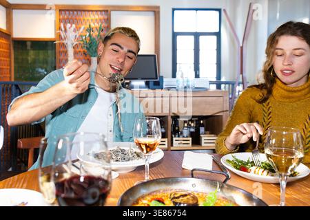 Les amis partagent un repas méditerranéen dans un restaurant confortable, dégustant paella et vin à une table en bois. Un homme sert des nouilles tandis qu'une femme mange de la salade, Re Banque D'Images