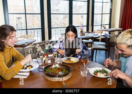 Trois amis apprécient la cuisine méditerranéenne dans un restaurant lumineux, partageant paella et fruits de mer frais tout en discutant et riant autour du vin dans un l détendu Banque D'Images