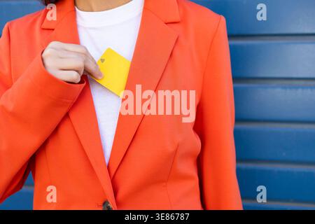 Une femme d'affaires dans un blazer orange vif met une carte de crédit jaune à l'intérieur de son blazer, symbolisant les transactions financières et les affaires Banque D'Images