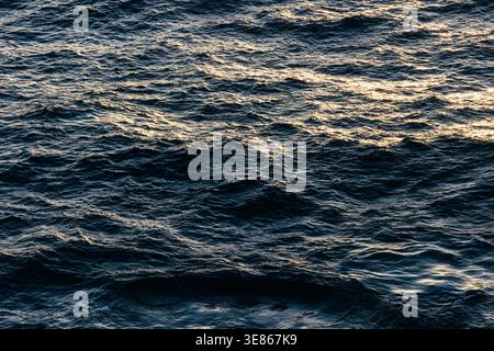Vue rapprochée de l'eau sombre de l'océan avec vagues roulantes et texture ondulante, éclairée par les reflets chauds de la lumière du soleil. Fond de paysage marin naturel avec mouvement a Banque D'Images