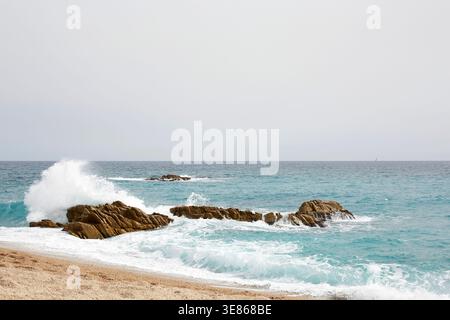 Paysage marin pittoresque d'eau turquoise et de vagues mousseuses s'écrasant contre des rochers accidentés près d'une plage de sable, sous un ciel pâle. Paysage côtier naturel avec Banque D'Images