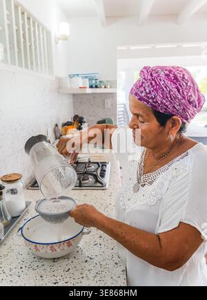 Une femme dans un foulard vibrant prépare du lait de coco frais dans une cuisine tropicale des Caraïbes. Le processus consiste à filtrer le mélange de noix de coco mélangé pour aut Banque D'Images