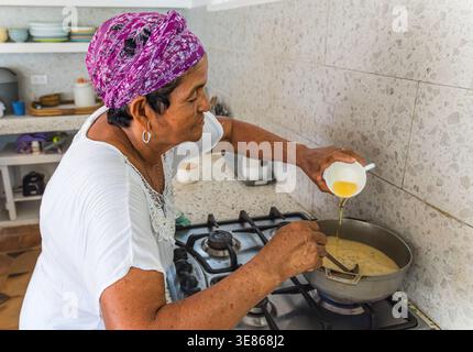 Une femme âgée portant un foulard coloré prépare habilement un plat traditionnel des Caraïbes dans une cuisine à la maison, versant des ingrédients dans une casserole Banque D'Images