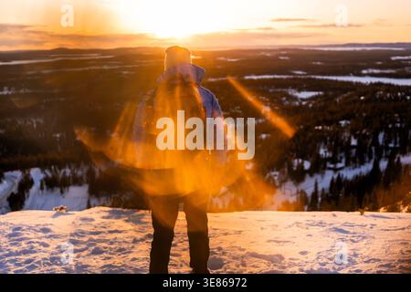Randonneur rétro-éclairé se tient sur une colline enneigée au coucher du soleil en Finlande, surplombant une vaste forêt et des lacs gelés, avec une lumière chaude du soleil créant une nature sereine Banque D'Images