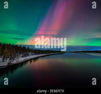 aurores boréales vibrantes en arcs verts et magenta à travers un ciel étoilé, luisant au-dessus d'une rivière calme avec des berges forestières enneigées dans l'hiver finlandais Banque D'Images