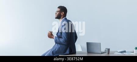 Homme en costume s'assoit sur le bureau tout en tenant la tasse dans le bureau moderne pendant les heures de travail Banque D'Images