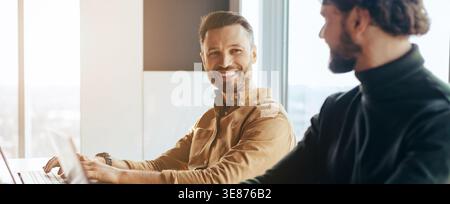 Deux hommes assis dans un bureau moderne souriant et partageant un moment pendant une journée de travail Banque D'Images