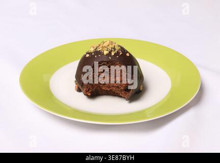 Un gâteau boule de chocolat à moitié mangé garni de pistaches écrasées servi sur une assiette verte et blanche sur une nappe blanche. Banque D'Images