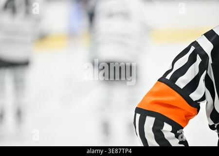 Close-up du maillot rayé et du brassard orange d'un arbitre de hockey sur glace, avec des joueurs flous patinant sur la glace en arrière-plan. Banque D'Images