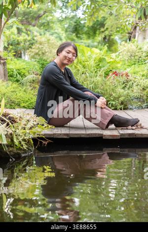 Femme LGBTQ assise près d'un étang dans un jardin luxuriant portant des lunettes détendues pose lumière naturelle cadre extérieur paisible reflétant une identité calme et inclusive Banque D'Images