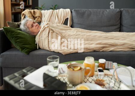 Femme caucasienne senior allongée sur un canapé sous une couverture, se reposant pendant la maladie avec des médicaments et du thé à proximité. moyen long shot, publicités de soins de santé adaptées, campagnes de saison froide, contenu de récupération à domicile Banque D'Images