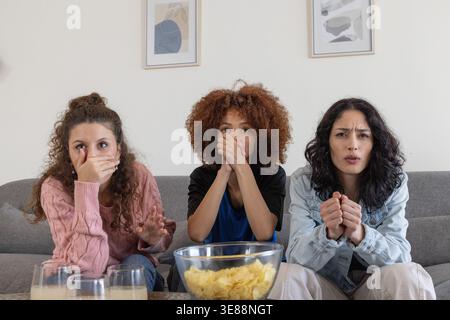 Diverses amies féminines assises sur un canapé gris dans le salon réagissant avec ténacité avec un bol de puces Banque D'Images