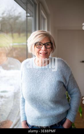Femme âgée souriante regardant par la fenêtre à la maison. Banque D'Images