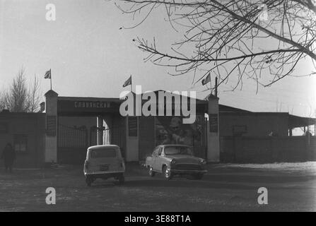 Sloviansk, 1975. Vue sur l'entrée principale de l'usine de transformation de viande locale. La porte solide arbore des drapeaux patriotiques et une grande affiche de propagande idéologique représentant Vladimir Lénine aux côtés de travailleurs enthousiastes. Deux voitures de tourisme soviétiques classiques sont garées directement devant les portes métalliques fermées par une journée d'hiver brillante et froide. Cette captivante photographie d'archives historiques en noir et blanc documente efficacement les installations industrielles, l'histoire de l'automobile ancienne et les routines civiques quotidiennes dans un Donbass paisible. Banque D'Images