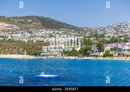 Ville côtière ensoleillée de Kalkan avec plage de sable, stations balnéaires et maisons blanches à flanc de colline surplombant la mer bleu profond calme, palmiers et créatine de bateaux Banque D'Images