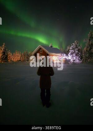 Un spectacle fascinant de aurores boréales danse dans le ciel nocturne à Akaslompolo, en Laponie. Un personnage se dresse en admiration, entouré par un pays des merveilles hivernales, avec une cabine chaude qui brille à proximité. Banque D'Images