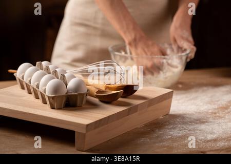 Mains pétrissant la pâte dans un bol en verre, préparant des produits de boulangerie faits maison, des œufs frais et des ustensiles de cuisson sur une planche de bois Banque D'Images