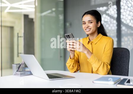 Jeune femme professionnelle souriant à la caméra tout en travaillant dans un bureau moderne, utilisant son smartphone et ayant un ordinateur portable ouvert sur le bureau pour la communication professionnelle et les tâches numériques Banque D'Images