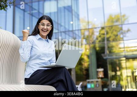 Jeune femme d'affaires souriant joyeusement, montrant un geste de pompe du poing, et tenant un ordinateur portable tout en étant assise sur un banc à l'extérieur d'un immeuble de bureaux en verre moderne, célébrant le succès des affaires Banque D'Images